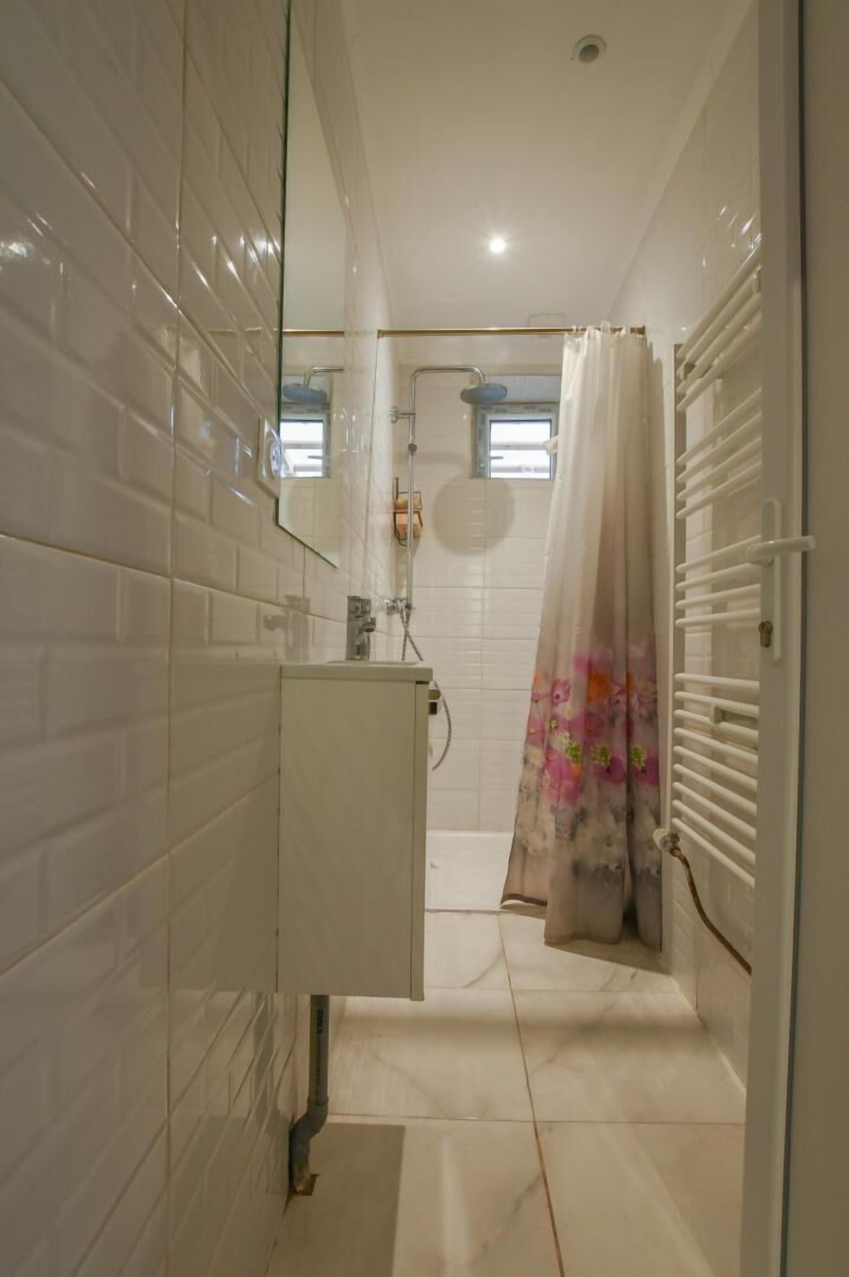 Une salle de bain avec des murs carrelés de blanc, un lavabo blanc et une douche agrémentée d’un rideau à motifs floraux.