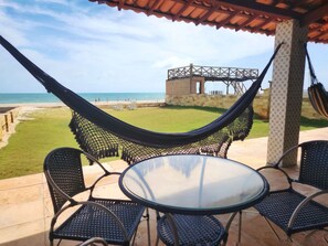 A patio with a glass table and chairs, a hammock, and a view of the beach and a stone structure. - Casa P na Areia com Piscina e Ar em Morro Branco (Beberibe)