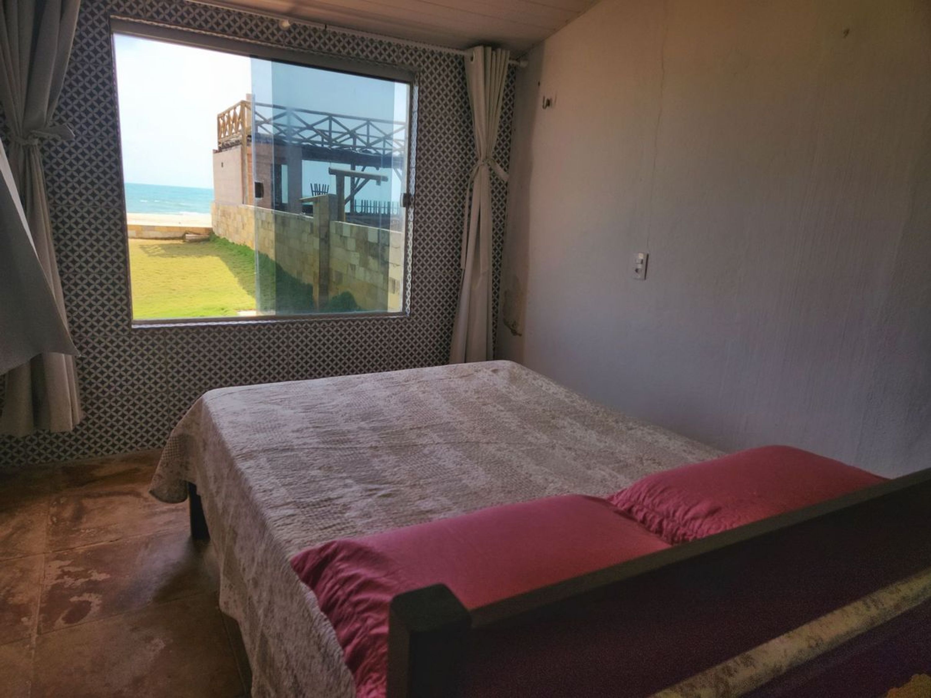En säng med en röd filt i ett rum med ett stort fönster med utsikt över en strand och en byggnad.