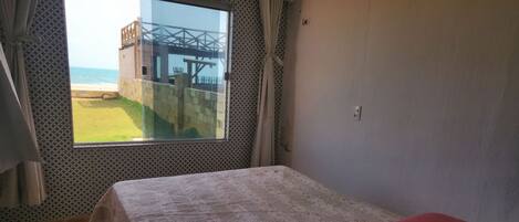 Una cama individual con una manta roja en una habitación con un ventanal grande que da a una playa y un edificio.