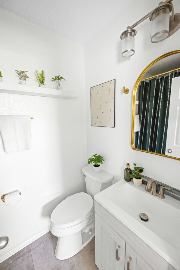 Un baño con inodoro blanco, lavamanos con grifo dorado y espejo con marco dorado. Hay plantas en el estante sobre el inodoro y un cuadro enmarcado en la pared.