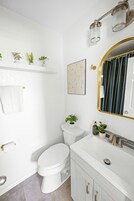 A bathroom with a white toilet, a sink with a gold faucet, and a mirror with a gold frame. There are plants on the shelf above the toilet and a framed picture on the wall.