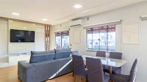 A modern interior with a grey sofa, a dining table with chairs, and large windows offering a view of buildings outside. - Apt With Barbecue 200 m From the Beach Ifa0054 (Penha)