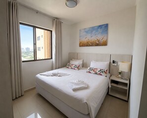 Ein Hotelzimmer mit einem großen Bett, einem Weizenbild an der Wand, einem Nachttisch mit Lampe und Blick auf Gebäude durch das Fenster.