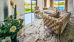 Ein großzügiger Wohnraum mit einer bequemen Couch, einem Couchtisch und einem großen Fenster mit Blick auf einen Swimmingpool und Grünflächen.