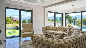 Ein geräumiges Wohnzimmer mit einer großen beigen Couch, einem Sessel und einem Glastisch. Große Fenster bieten Blick auf einen Swimmingpool und Berge.