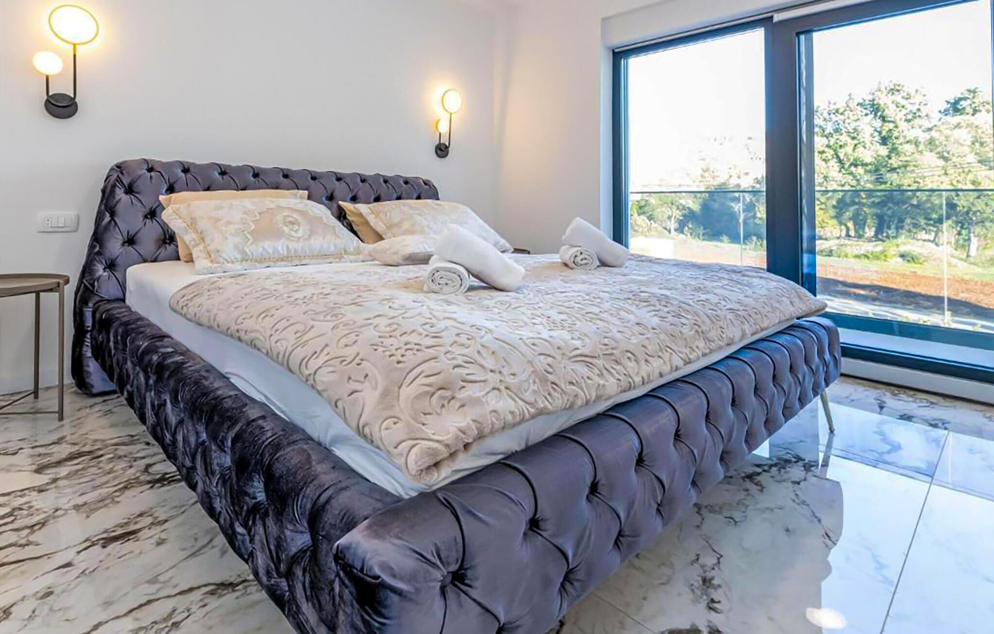 A bedroom with a large bed, two wall-mounted lights, and a view of greenery outside.