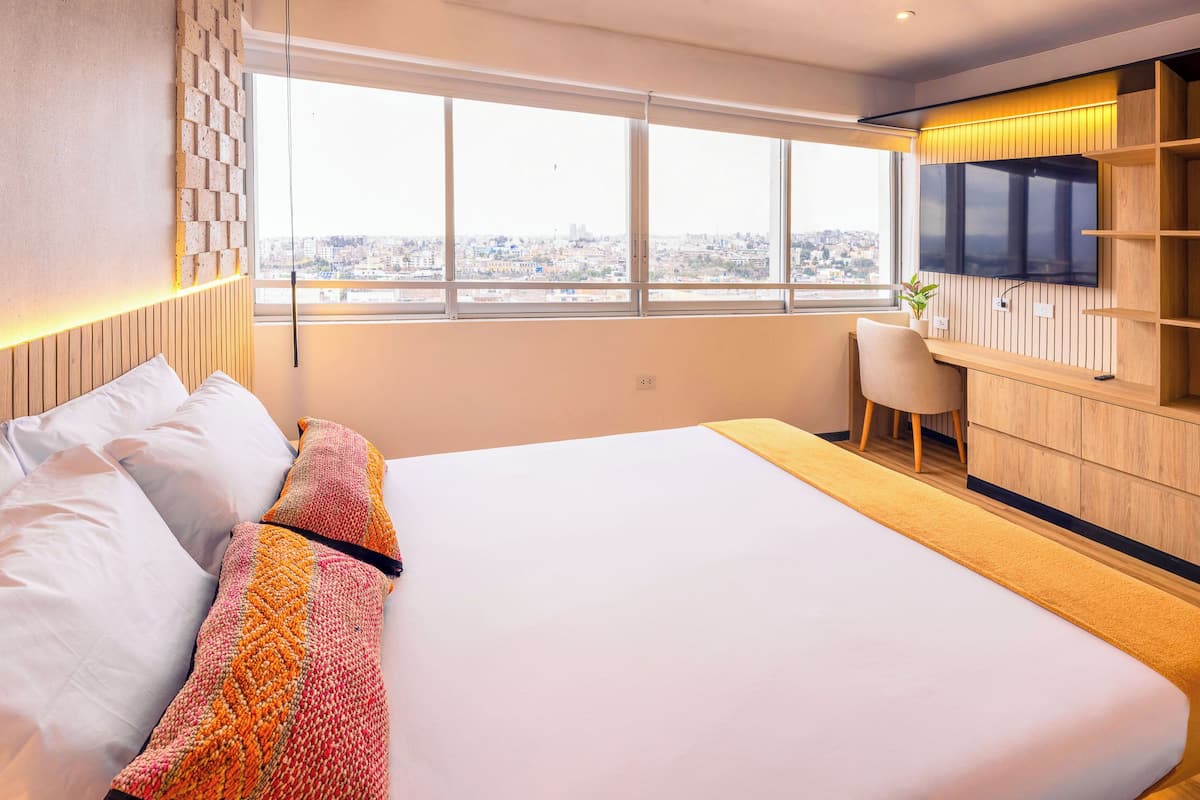 A modern hotel room with a large bed, a desk, a TV, and a city view.