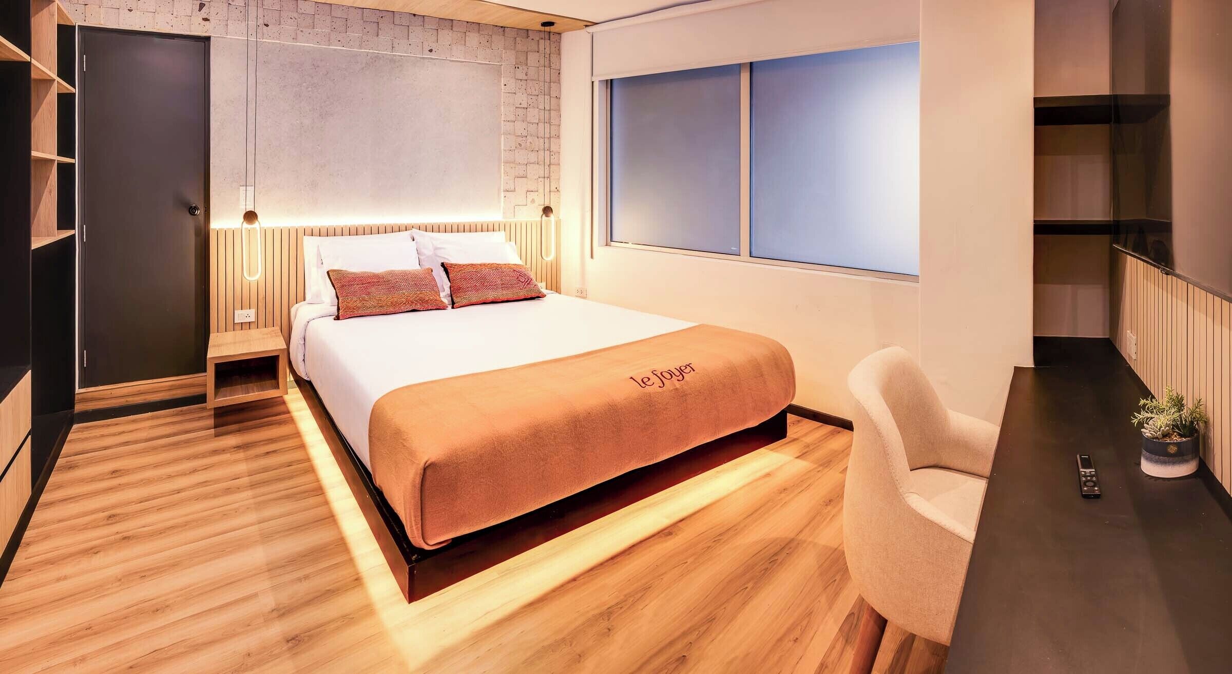 Une chambre d’hôtel moderne avec un plancher en bois, un lit recouvert d’une couverture beige, une table de chevet, une lampe, une chaise et une fenêtre avec des stores.