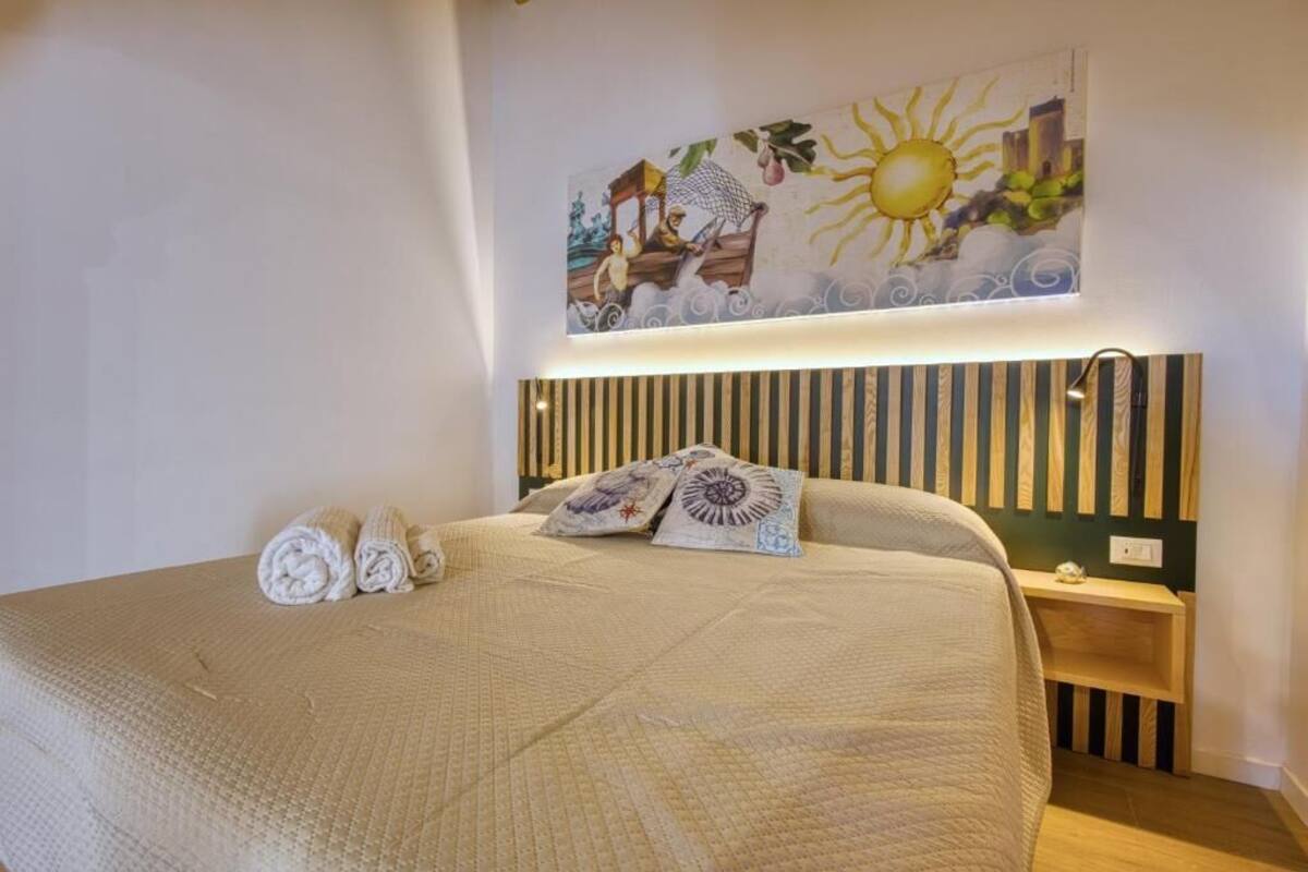 A neatly made bed with rolled towels, a decorative pillow, and a wall-mounted light fixture.