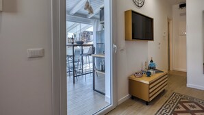 A modern interior with a sliding glass door leading to an outdoor area, a wooden console table with decorative items, a mounted television, and a wall clock. - VFH La Rada Apartments (Porticello)