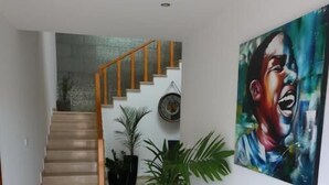 A hallway with a staircase, potted plants, and a large painting on the wall. - Lali's House (Tarrafal)