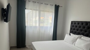 A hotel room with a large bed, a flat-screen TV mounted on the wall, and a view of the outdoors through sheer curtains. - Lali's House (Tarrafal)