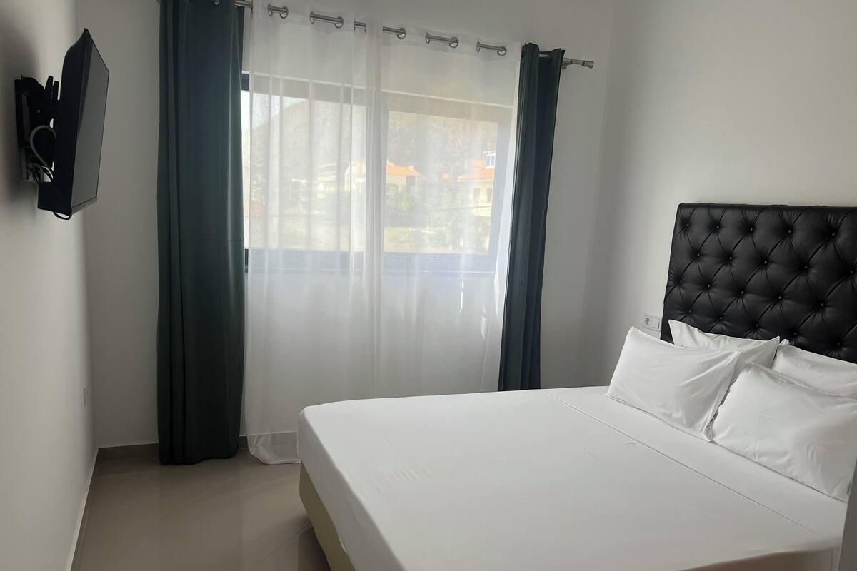 A hotel room with a large bed, a flat-screen TV mounted on the wall, and a view of the outdoors through sheer curtains.