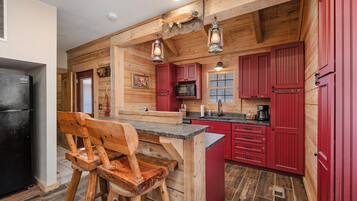 A rustic kitchen with wooden walls, a black refrigerator, red cabinets, and a wooden bar stool.