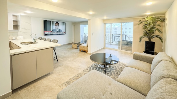 Un appartement moderne comprenant une cuisine, un coin repas et un salon. Il est équipé d’une grande télévision, d’un balcon avec vue et d’une plante en pot.