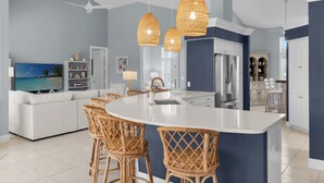 Een moderne keuken met een centraal eiland, witte kastjes en een groot eilandgootsteen. Er staan rieten stoelen rondom het eiland, een plafondventilator en hanglampen.