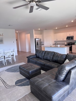 A modern living room with a black leather sectional sofa, a dining area with white chairs, and a kitchen with white cabinets and stainless steel appliances.