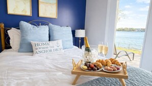 Una cama con una bandeja de bocadillos, una botella de champán y copas. Hay dos cuadros en la pared, un ventanal con vista al agua y una cama con un almohadón que dice 'HOME IS WHERE THE ANCHOR DROPS'.