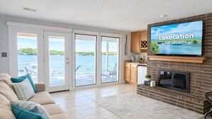 Una sala de estar con una televisión que muestra 'Lakecation vacations', un sofá con cojines color turquesa, una chimenea y vista al agua a través de amplias puertas correderas de vidrio.