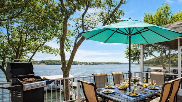 Una terraza al aire libre con una parrilla, una mesa de comedor, sillas y vistas a un lago.