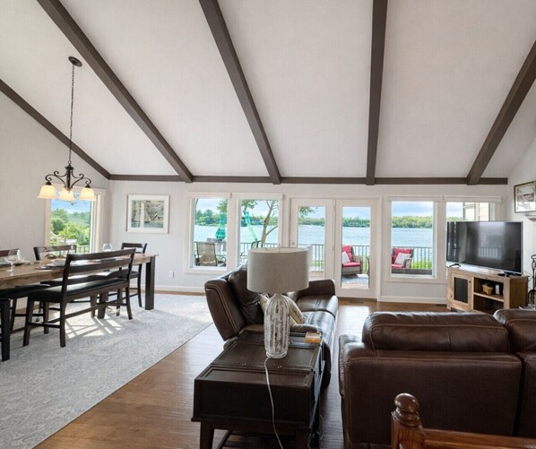A spacious living area with a dining table, leather sofas, a TV, and large windows overlooking a body of water.