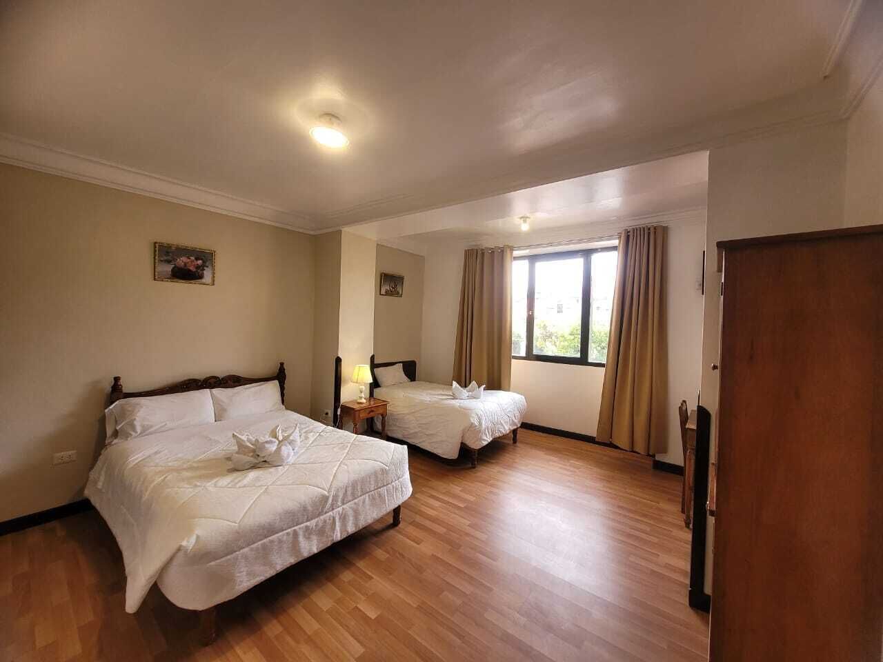 A hotel room with two beds, wooden flooring, a window with curtains, and a door.