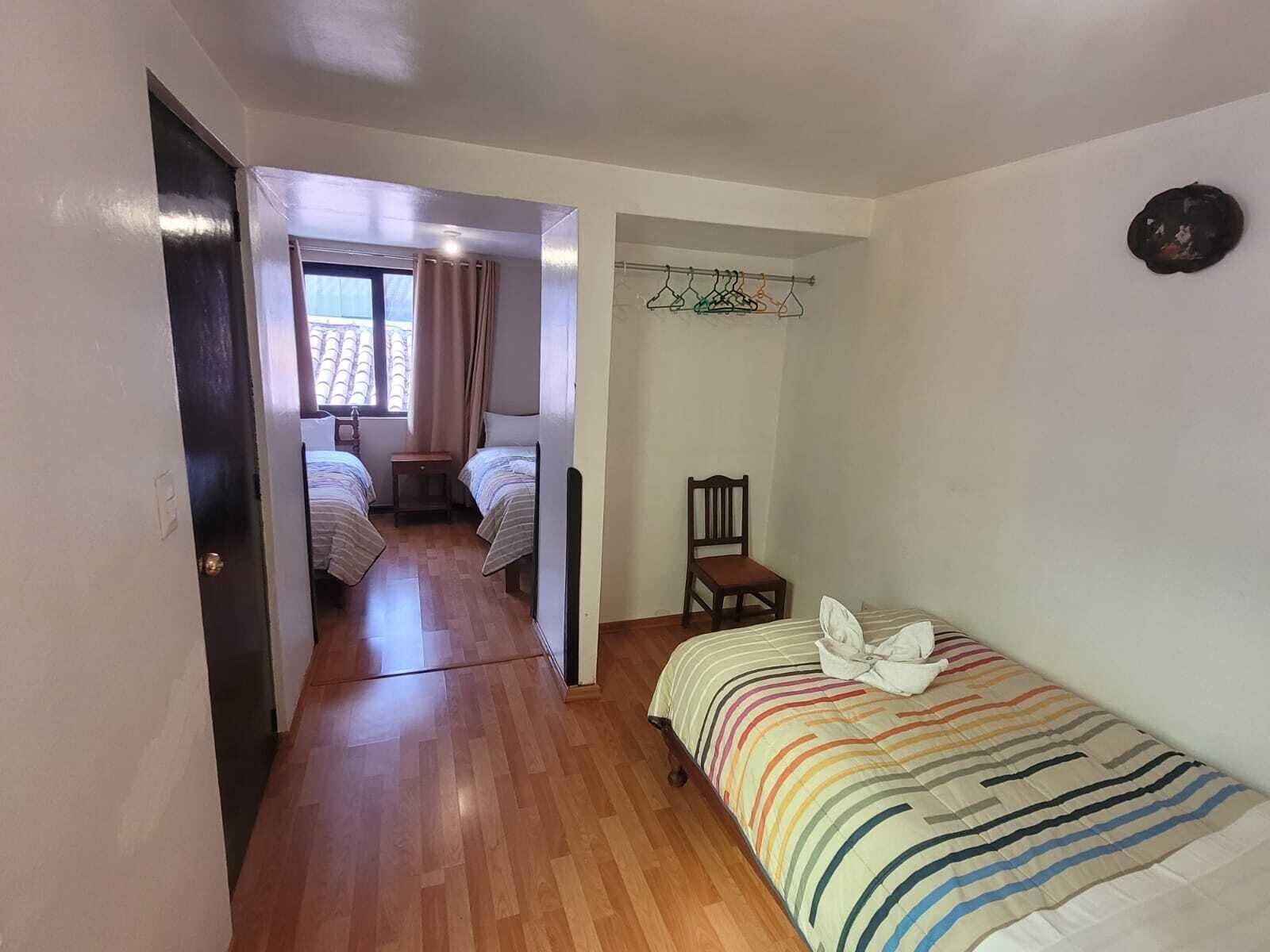 A room with a wooden floor, a bed with a striped bedspread, a chair, a coat rack with clothes, and a window with curtains.
