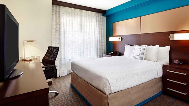Quarto de hotel com cama, escrivaninha, televisão, cadeira e mesa de cabeceira.