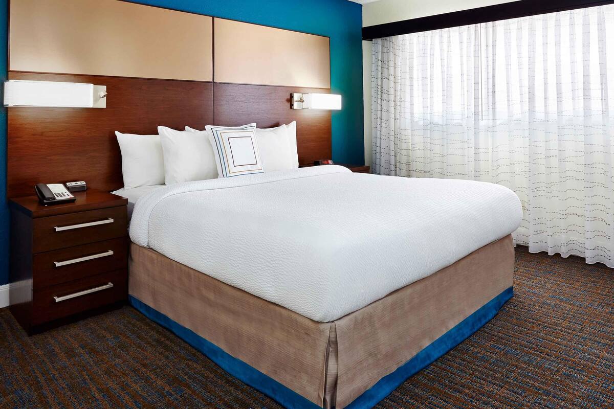 A hotel room with a large bed, a wooden headboard, a nightstand with a phone, and a window with sheer curtains.
