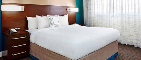 A hotel room with a large bed, a wooden headboard, a nightstand with a phone, and a window with sheer curtains.