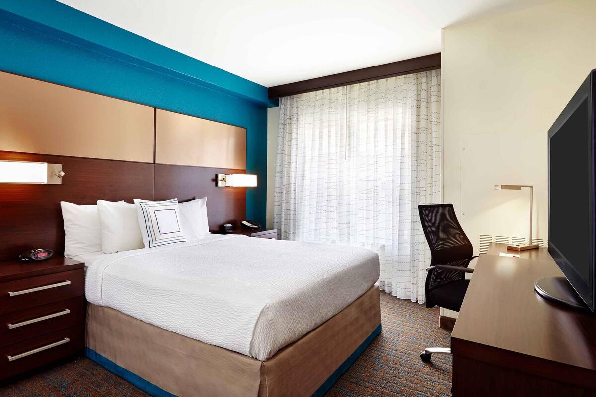 A hotel room with a large bed, a desk with a television, a chair, a nightstand, and a window with curtains.