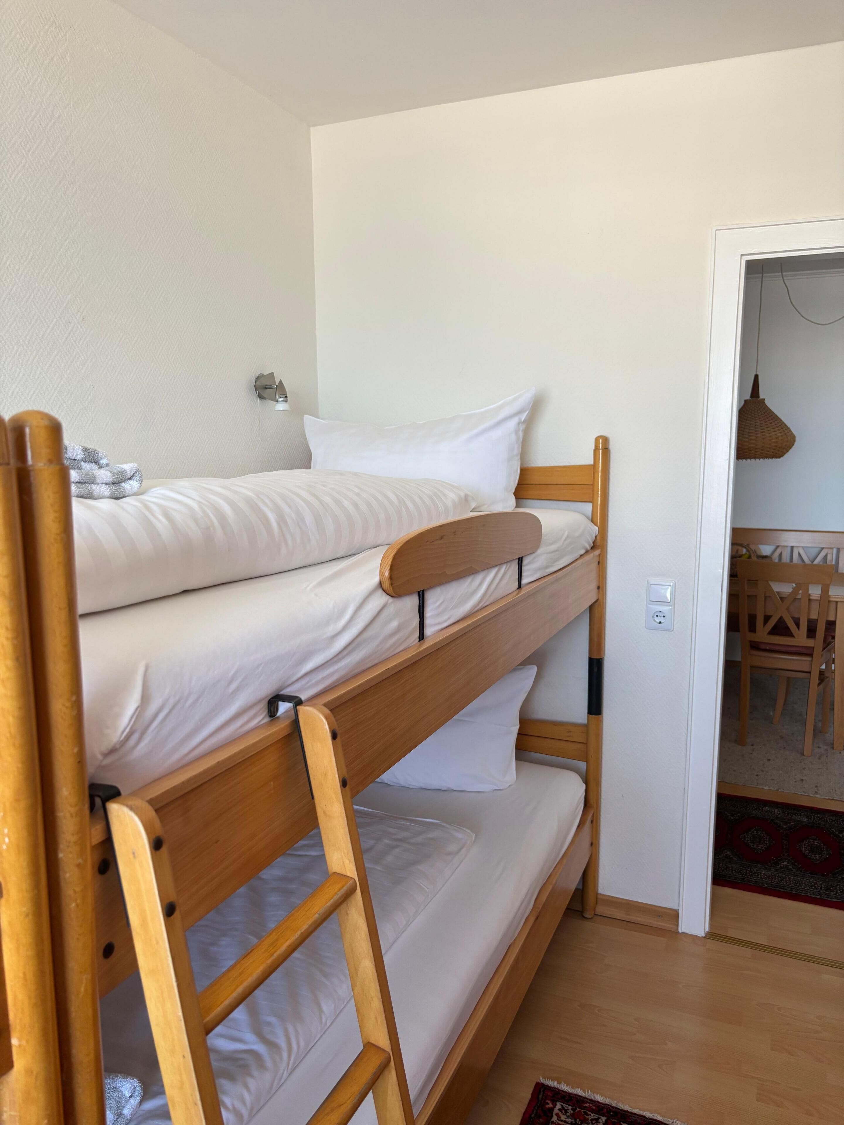 Una cama litera con ropa de cama blanca, un cabecero de madera y una pequeña mesa con sillas en la habitación contigua.