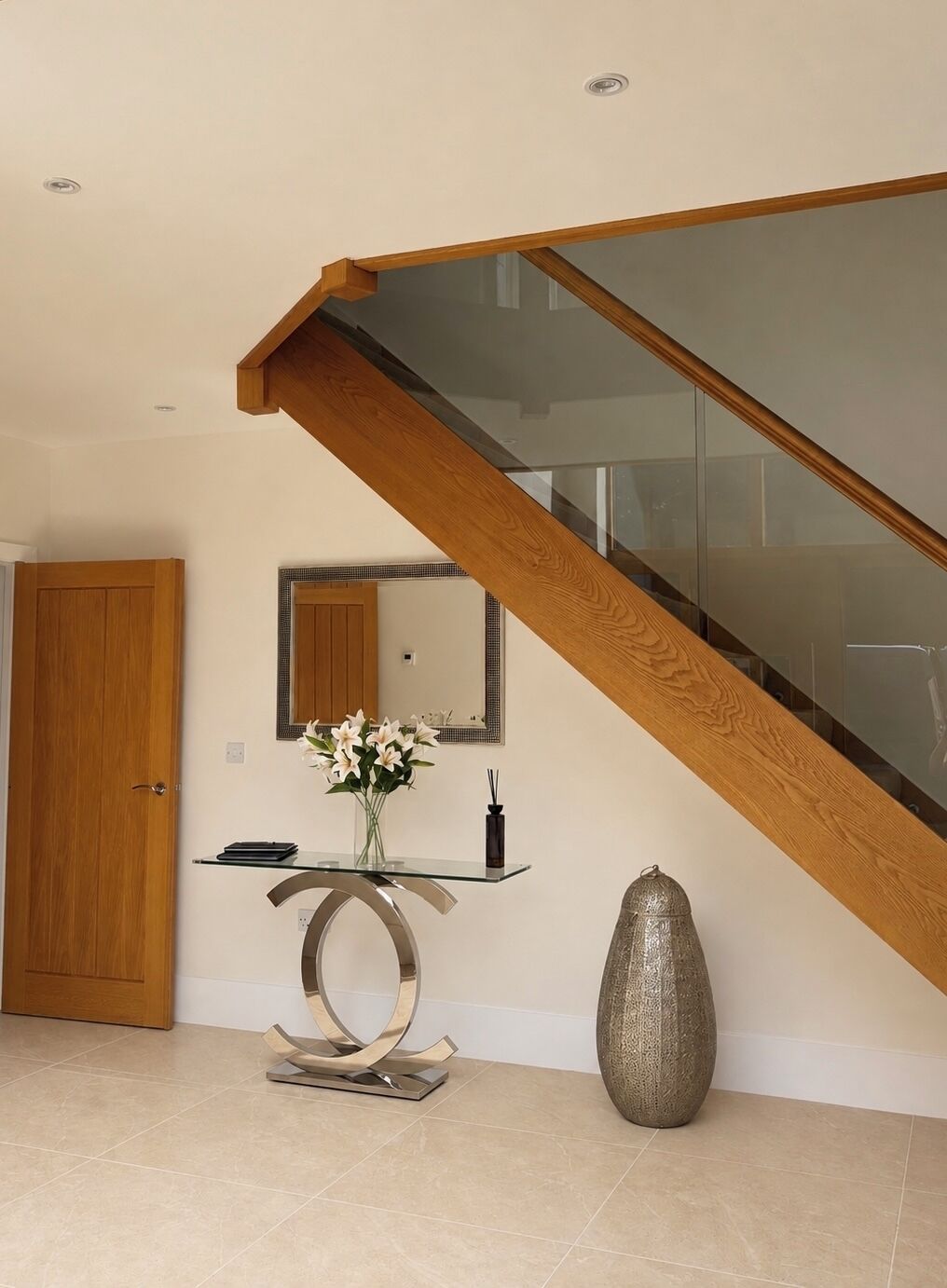 A modern interior with a wooden staircase, a glass railing, a wooden door, a glass console table with a vase of flowers, and a textured vase.