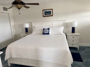 A neatly made bed with a quilted white comforter, two bedside tables with lamps, a ceiling fan, a framed picture on the wall, and a dark blue rug on the floor.