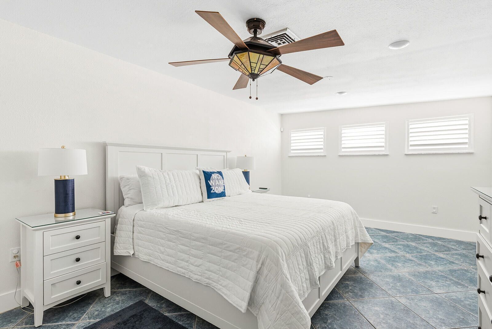 A bedroom with a bed, nightstand, lamp, ceiling fan, and windows.