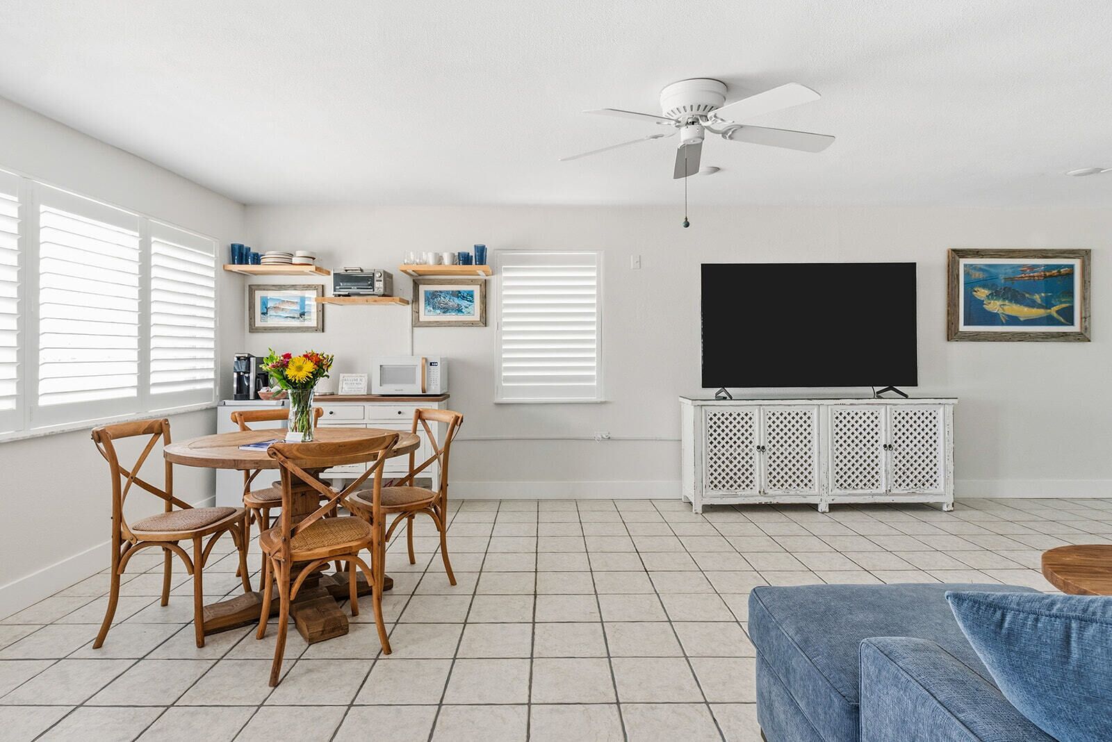 A living room with a dining area, white walls, a ceiling fan, a flat-screen TV, and a white cabinet.