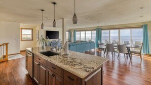 A spacious kitchen with granite countertops, wooden cabinets, and pendant lighting. A large island with a sink is central, and the room opens to a dining area with a view of the ocean. - The Salt Life - Oceanfront escape Hot tub (Lincoln City)