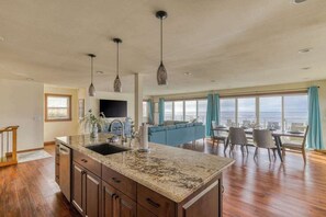 A spacious kitchen with granite countertops, wooden cabinets, and pendant lighting. A large island with a sink is central, and the room opens to a dining area with a view of the ocean. - The Salt Life - Oceanfront escape Hot tub (Lincoln City)