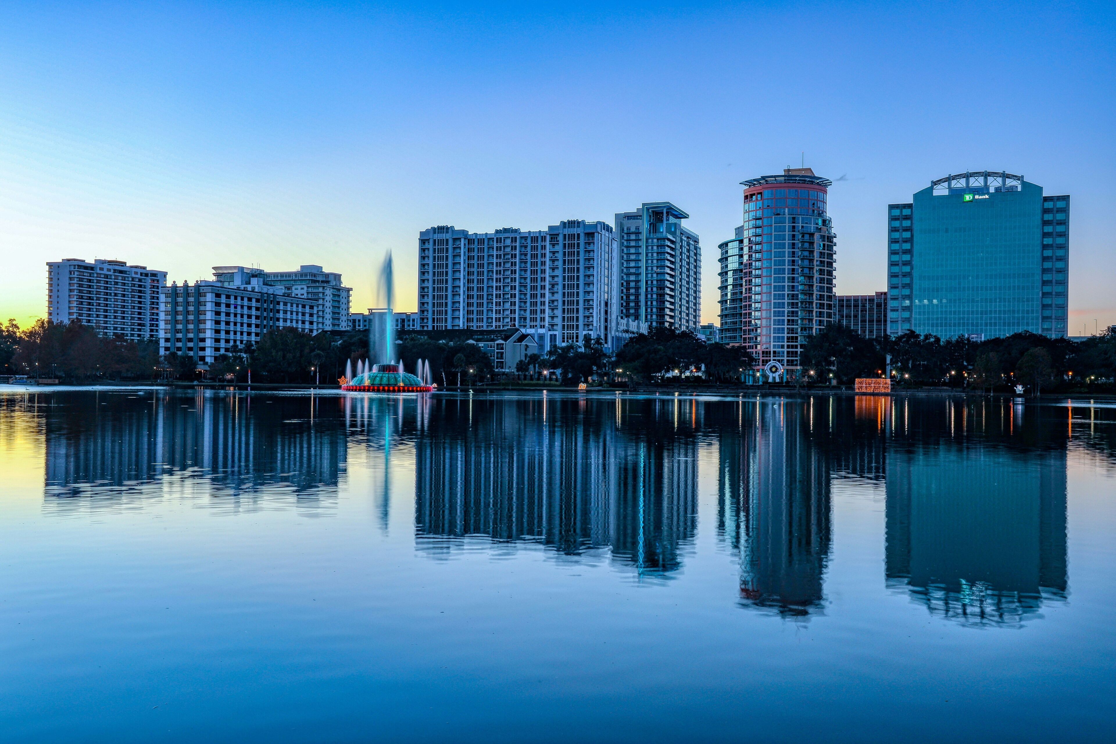 A city skyline with high-rise buildings, a body of water reflecting the structures, and a prominent tower with a dome-like structure in the foreground.