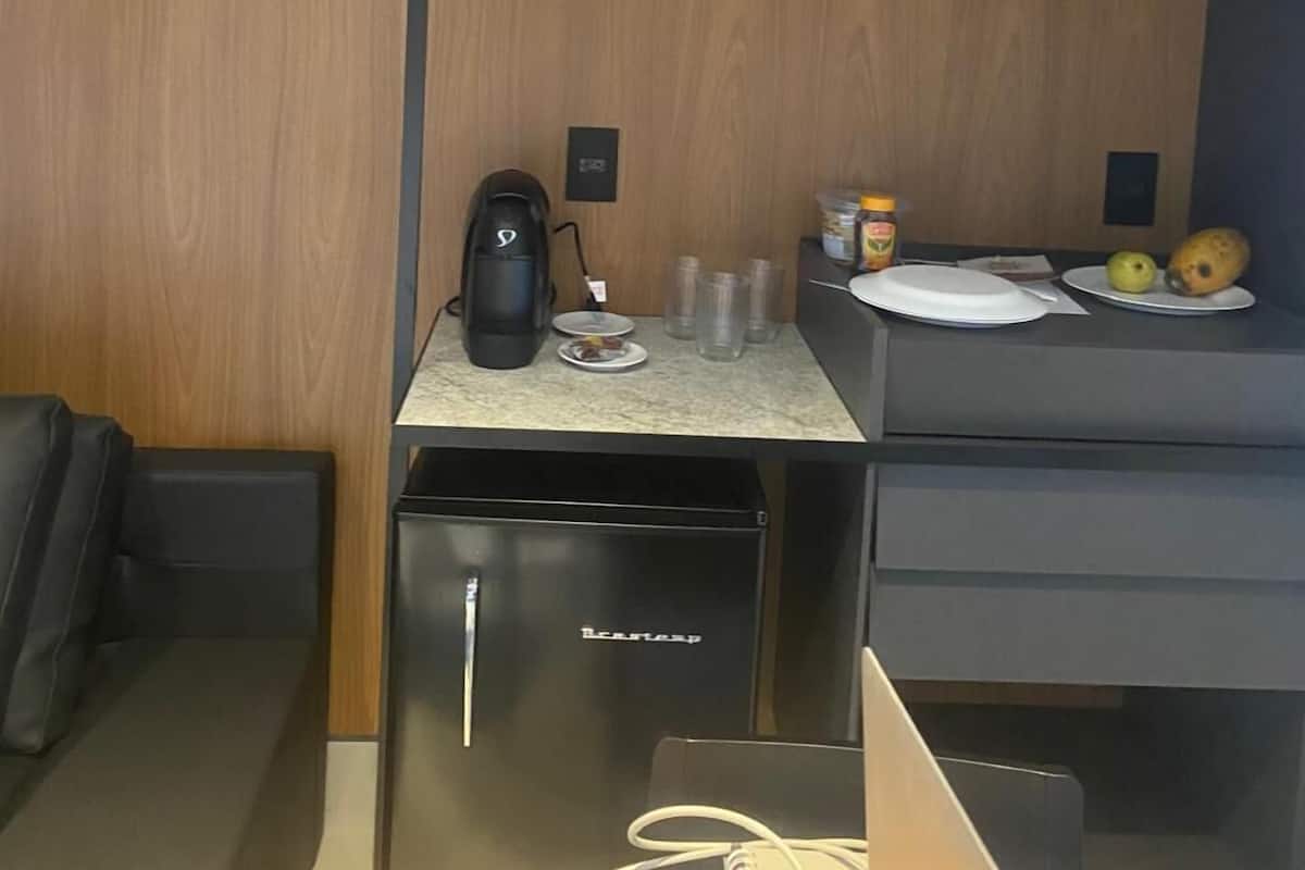 A compact kitchenette with a coffee maker, a small fridge, and a counter with a plate, glasses, and fruits.