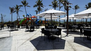 Un'area patio esterna con tavoli e sedie, palme e un'area giochi con scivoli.