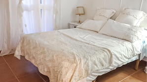 Un dormitorio con cama, cortinas blancas, una mesita de noche con lámpara y un velador.