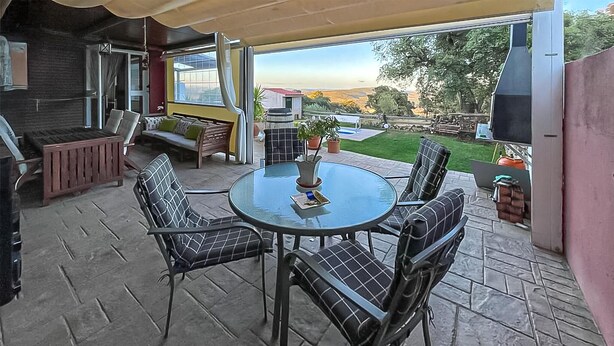 Un patio al aire libre con una mesa redonda de vidrio y cuatro sillas, una parrilla y vistas a un jardín y colinas al fondo.