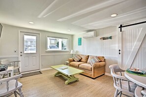Ett mysigt vardagsrum med ett grönt soffbord, en beige soffa och vita stolar. Det finns två fönster med utsikt över omgivningen, en dörr och en väggmonterad luftkonditioneringsanläggning.
