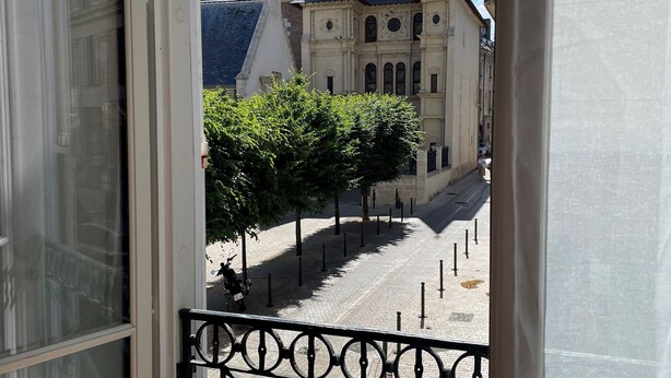 A view from a window overlooking a cobblestone street with historic buildings, a metal railing, and a clear blue sky.