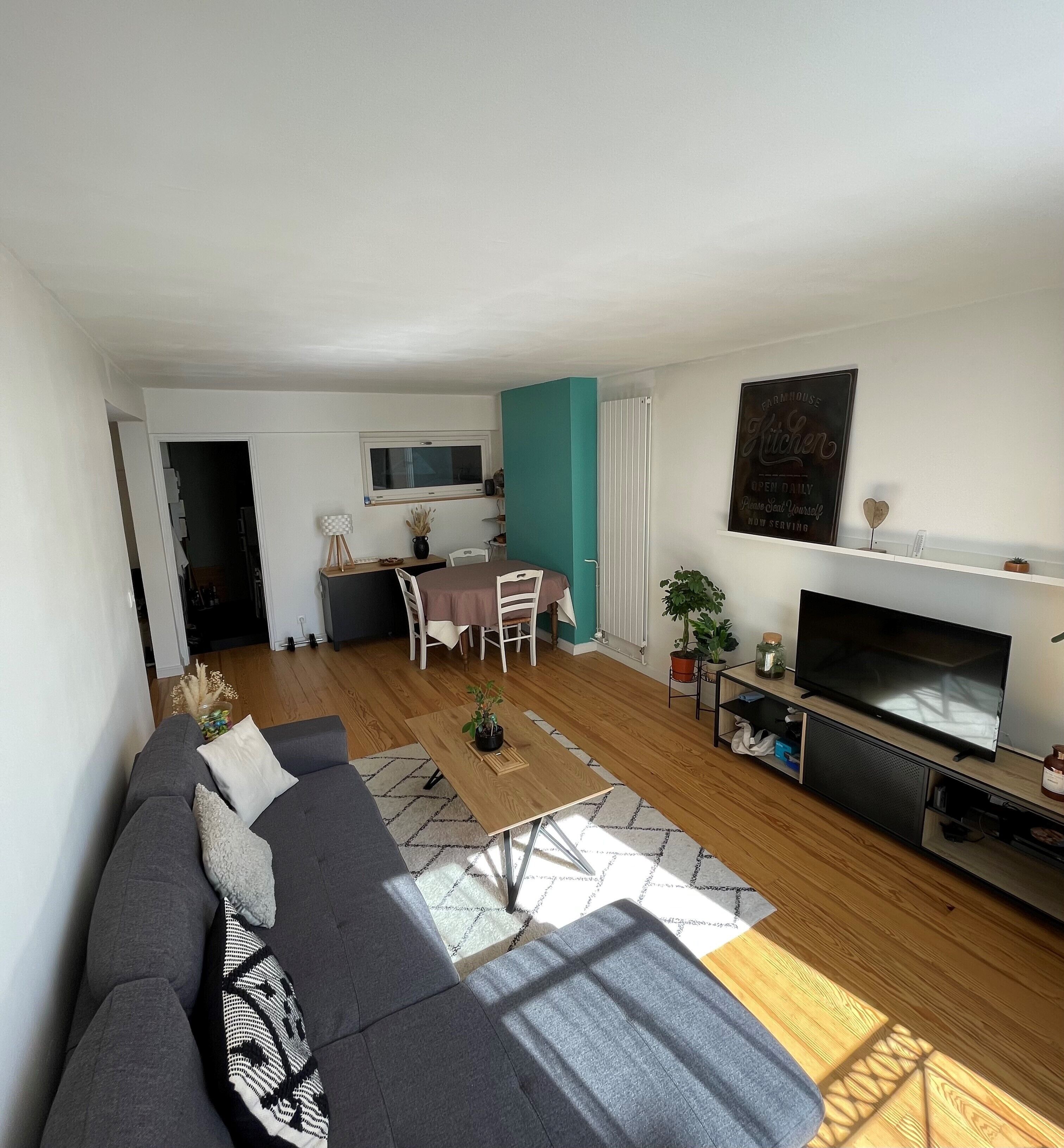 En moderne stue med en grå sofa, et spisebord med stole, et fjernsyn og et køkken i baggrunden.