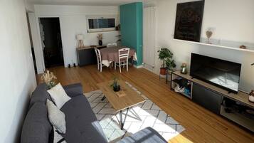 En moderne stue med en grå sofa, et spisebord med stole, et fjernsyn og et køkken i baggrunden.