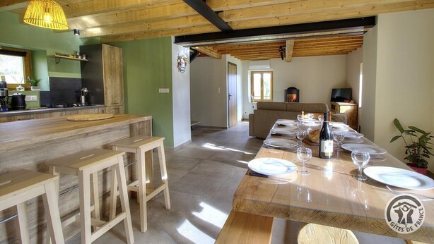 A rustic kitchen with wooden beams, a bar area with stools, and a dining table set for six with wine glasses and plates. - "Ensemble - MEYS - Gîte (MEYS)