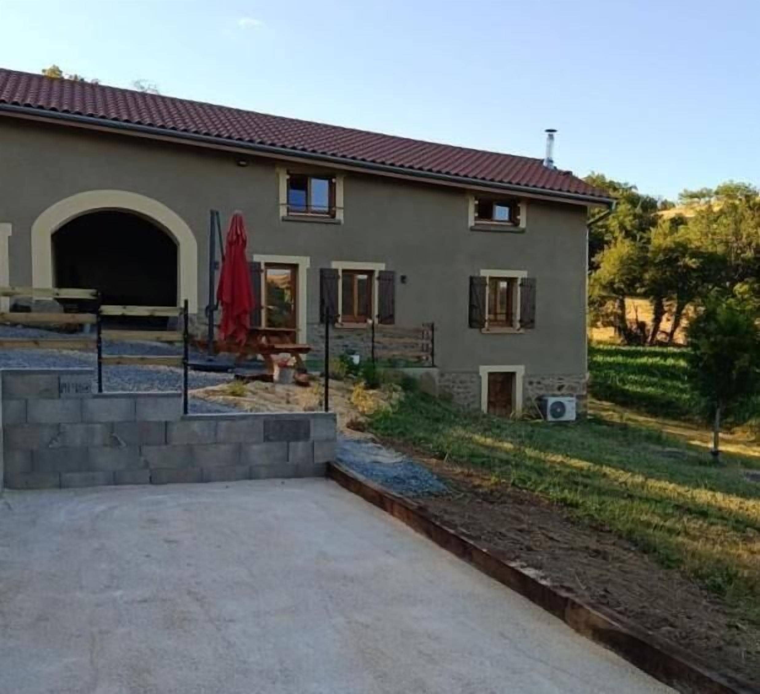 Une maison à deux étages avec une façade en pierre, un toit recouvert de tuiles rouges et une allée pavée menant à un patio abrité.